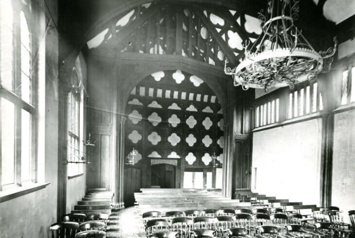 A black and white photograph of the Great Hall at Ordsall Hall with full of chairs. There is a chandelier hanging from the chunky wooden beams on the ceiling. The back wall is covered in a Quatrefoil pattern.