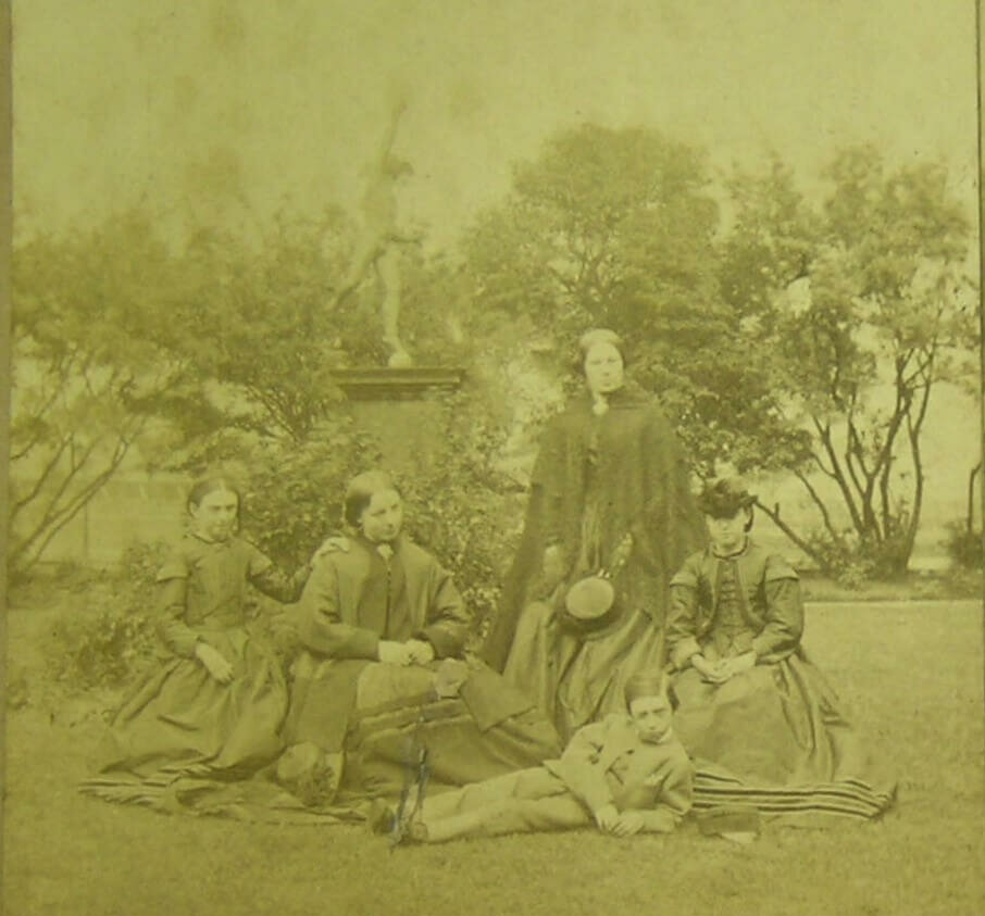 A greyscale photograph of three women and two children in old fashioned clothes posing for a photograph in front of a statue. The little boy reclines in the grass in front of the ladies.