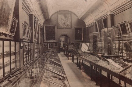 A black and white photo of an exhibition space. There are framed pictures on the walls and rows of display cases on the floor.