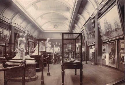 A black and white photograph of a gallery with a curved ceiling. There are paintings hanging on the walls, display cases and marble statues on plinths.