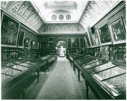 A black and white photo of a gallery that is full of display cases. There is a decorative curved ceiling and the walls are full of framed paintings.