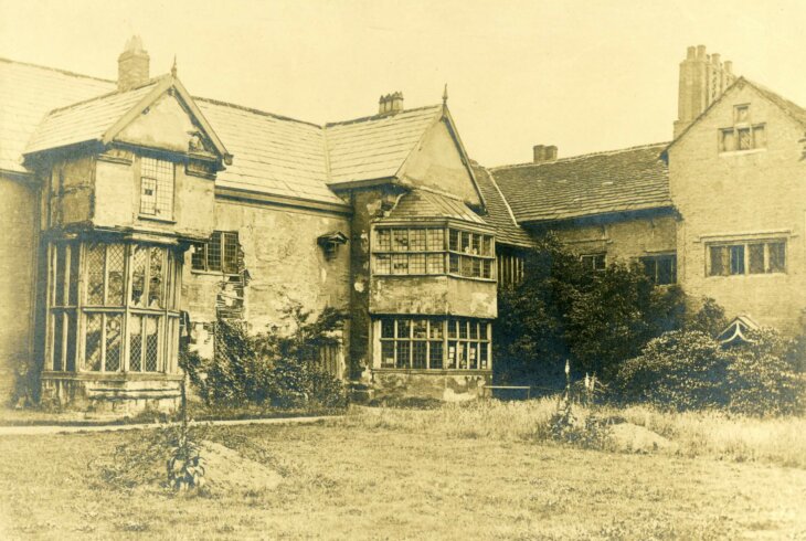 A greyscale photograph of Ordsall Hall looking run down. The large building has damaged windows and walls, and the grass is overgrown.