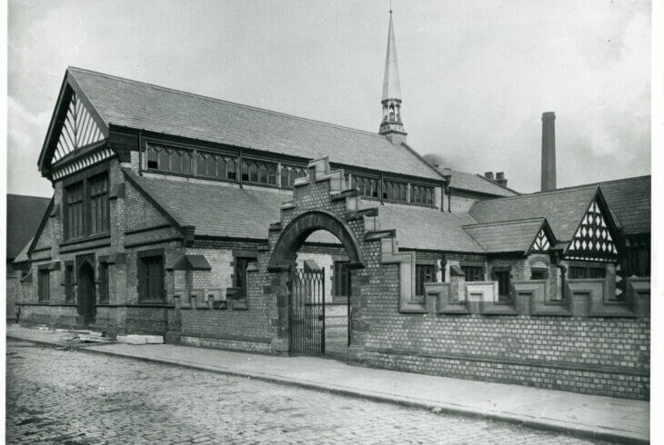 A black and white photograph of a large building with a Tudor roof. There is a brick wall with a decorative arch in front of the building.