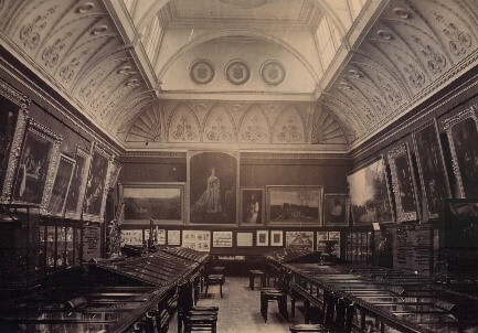 A black and white photograph of a gallery with a decorative, curved ceiling. The walls are full of framed paintings and there are many display cases on the floor.