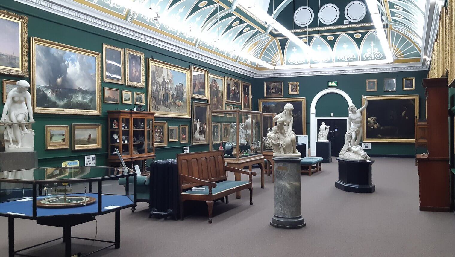 A gallery with dark green walls and a curved ceiling. There are framed paintings hanging on the walls and sculptures on plinths in the middle of the room.
