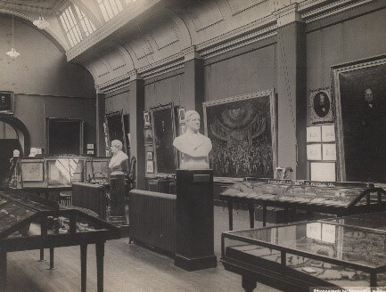 A black and white photograph of a gallery. Marble busts are on plinths in the middle of the room, surrounded by display cases. Framed paintings hang on the walls.