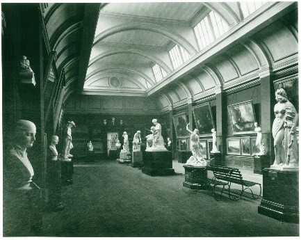 A black and white photo of a gallery with a high ceiling. The photo is old and looks green tinted. The room has paintings on the walls and statues on plinths.