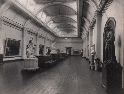 A black and white photo of a spacious gallery with a curved ceiling. The floors are empty apart from a few statues on plinths.