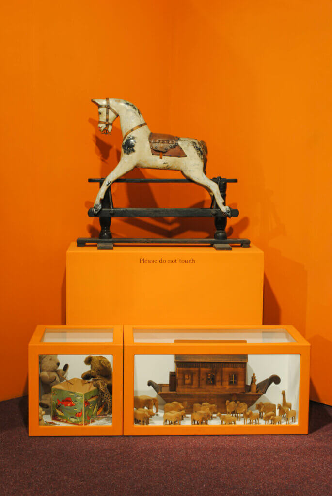 A white rocking horse on an orange plinth. In front of this are toys in display cases.