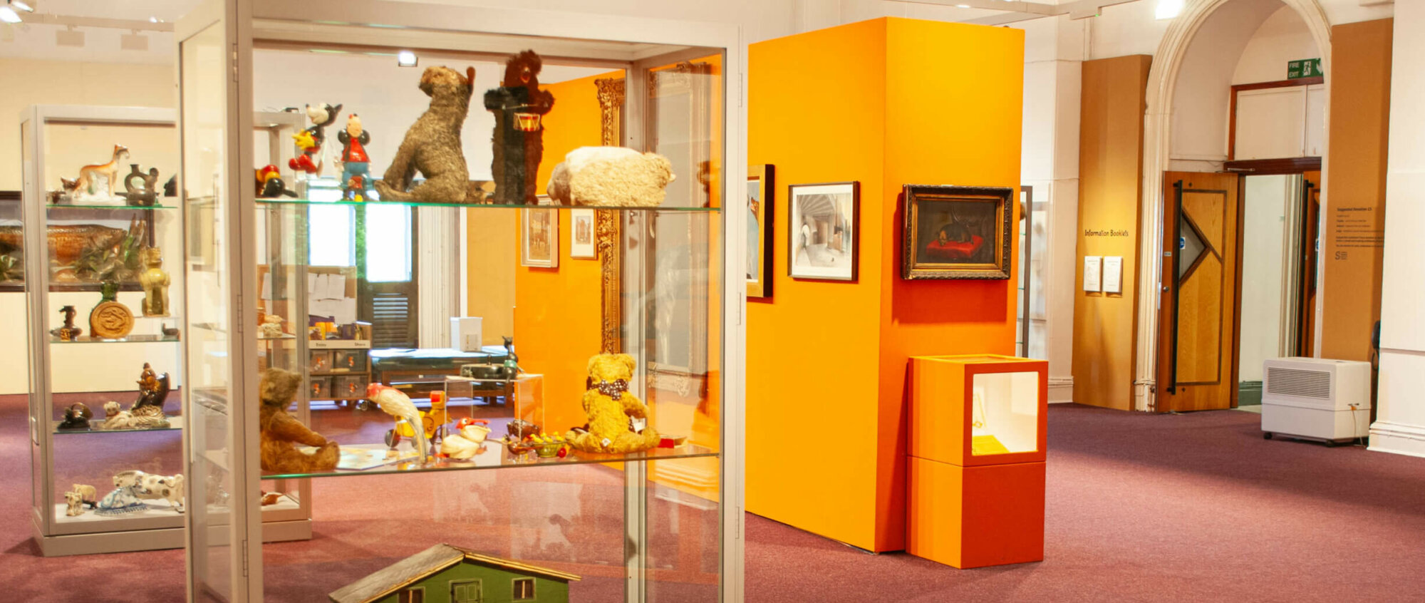A room with pink floor and yellowy orange walls. Art hangs on the walls and children's toys are displayed in cases.