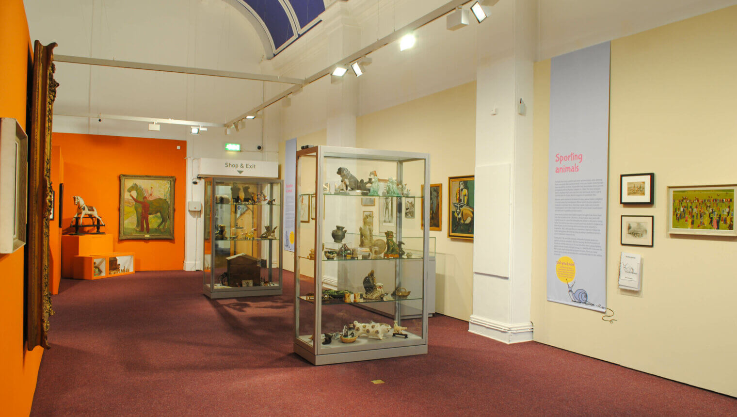A room with red carpets and walls painted cream or orange with framed paintings hanging on them. There are glass display cases containing sculptures of animals.