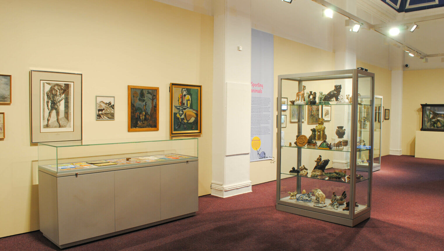 A room with red carpets and walls painted cream with framed paintings hanging on them. There are glass display cases containing sculptures of animals.
