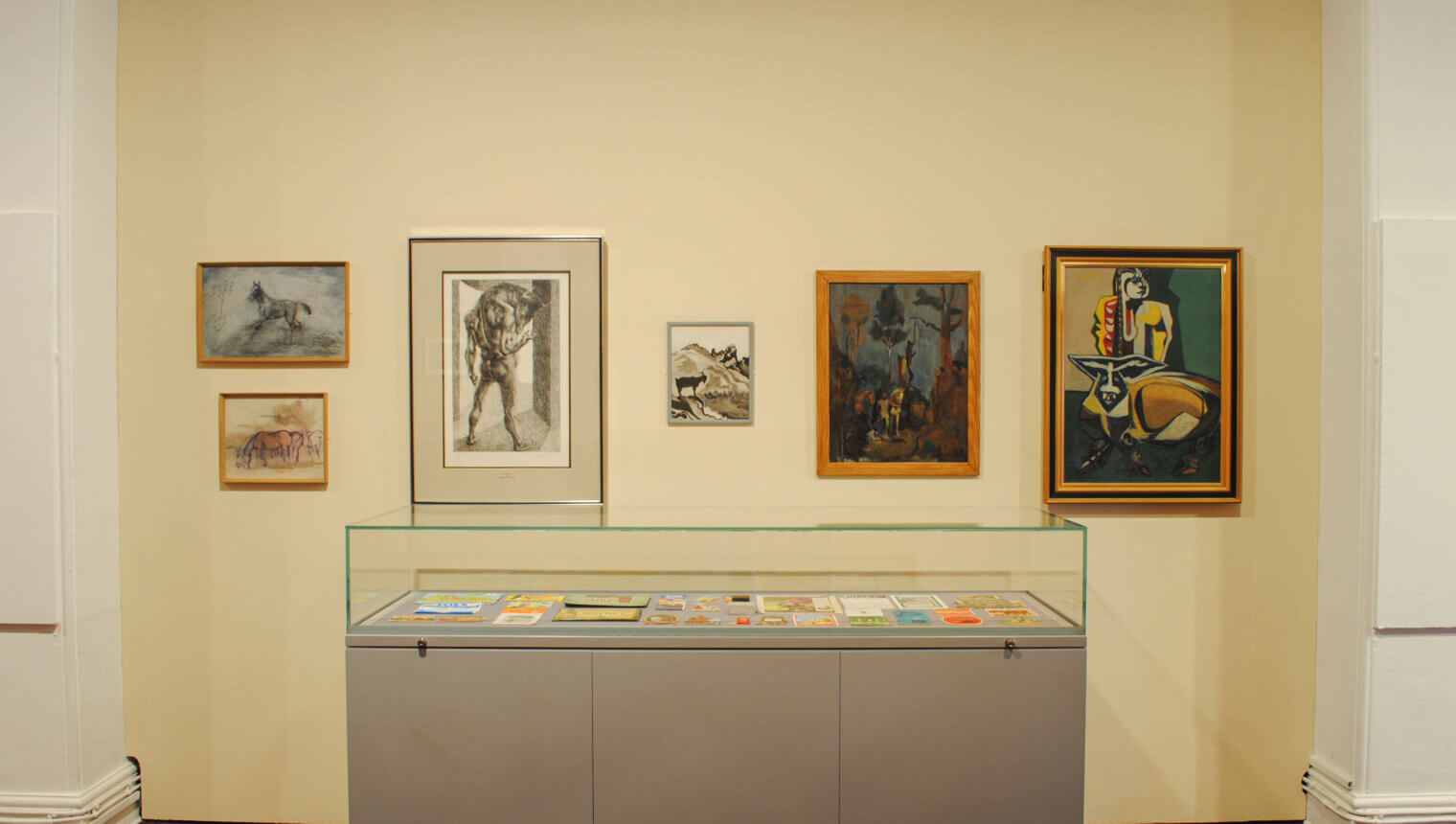 A cream wall with framed paintings hanging on it. There is a display case with paper works lying in.