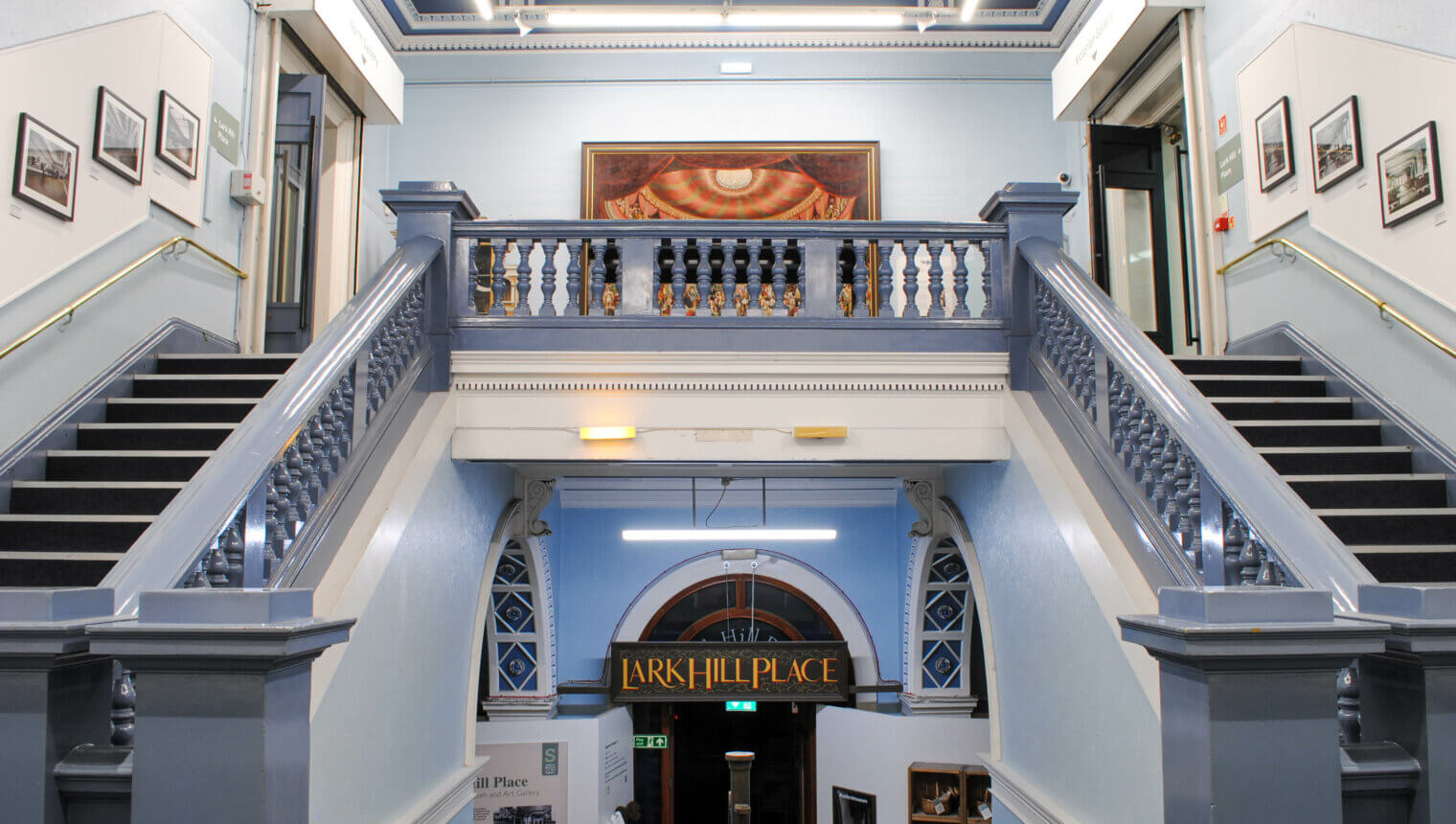 A grand double staircase with pale blue banisters. On the walls around it there are framed pictures.