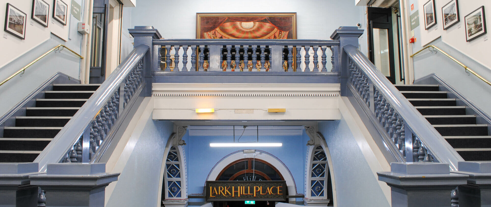 A grand double staircase with pale blue banisters. On the walls around it there are framed pictures.
