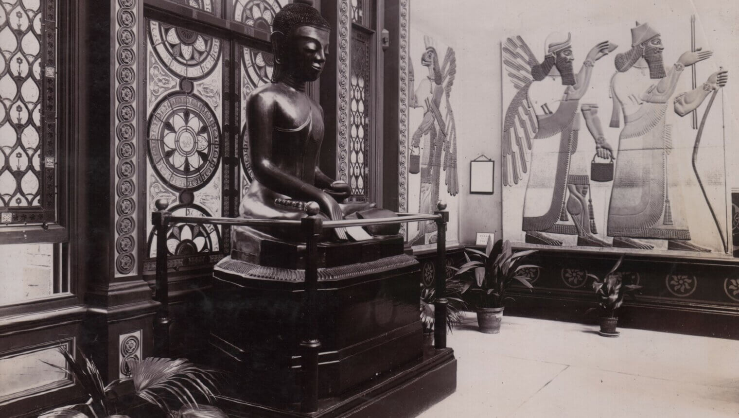 A statue of Buddha in front of a stain glass window. On the wall behind it are paintings of Egyptian gods.