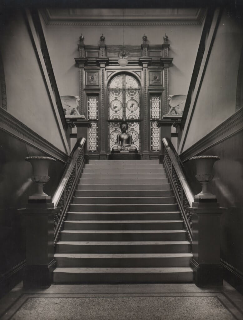 A flight of stairs with a statue of Buddha at the top sitting in front of a decorative window.