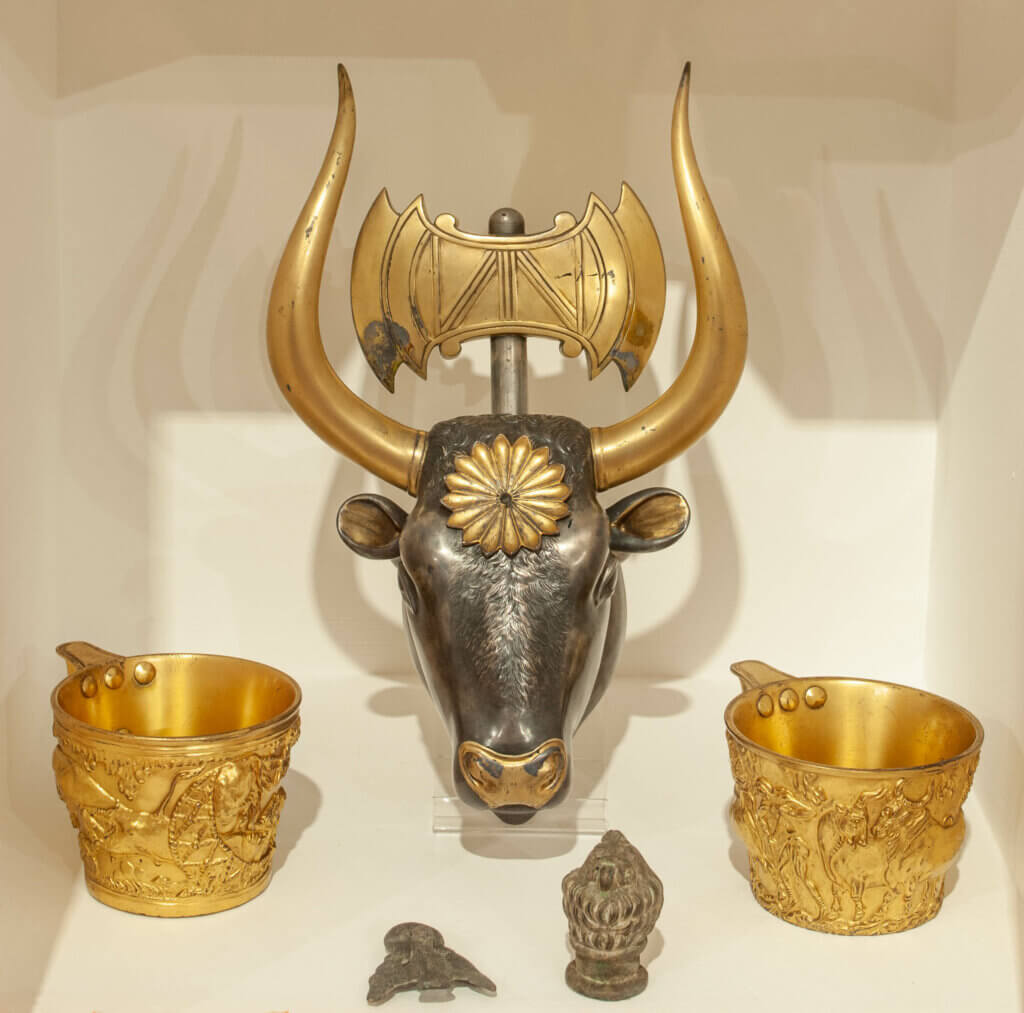 An array of ornate objects. Two golden cups with images of animals on rest on either side of a statue of a bull's head with horns. There is a hammer sticking out of the bull's head.