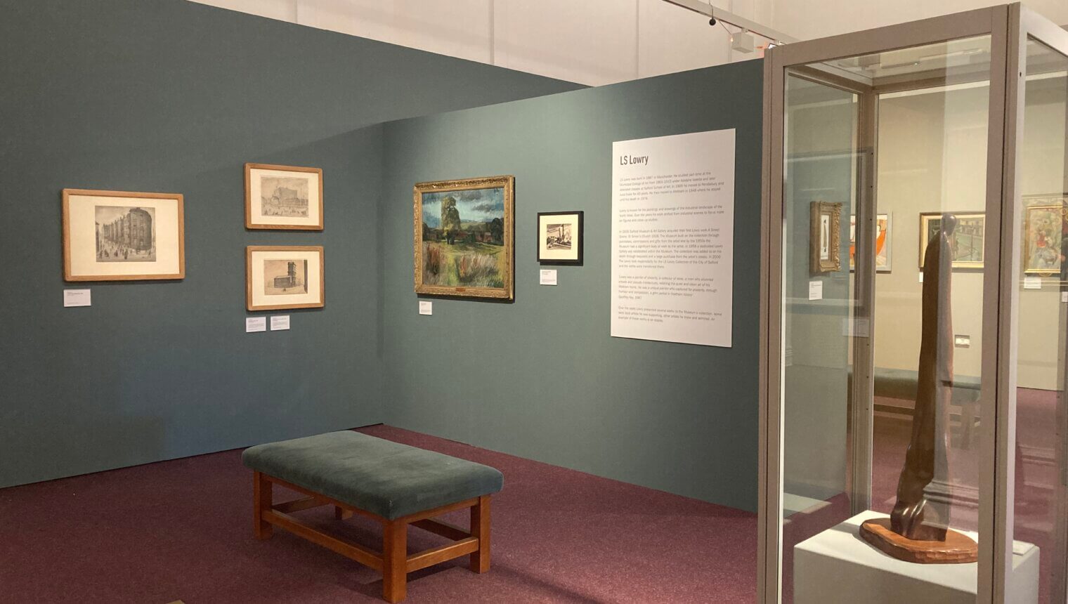 Blue walls with framed paintings on them. A label introducing the Lowry section. A sculpture in a case in the foreground.