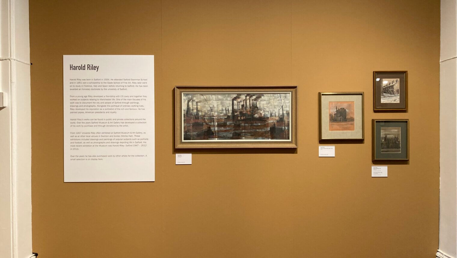A yellow wall with framed paintings hanging on it. A piece of writing introducing the Harold Riley section.