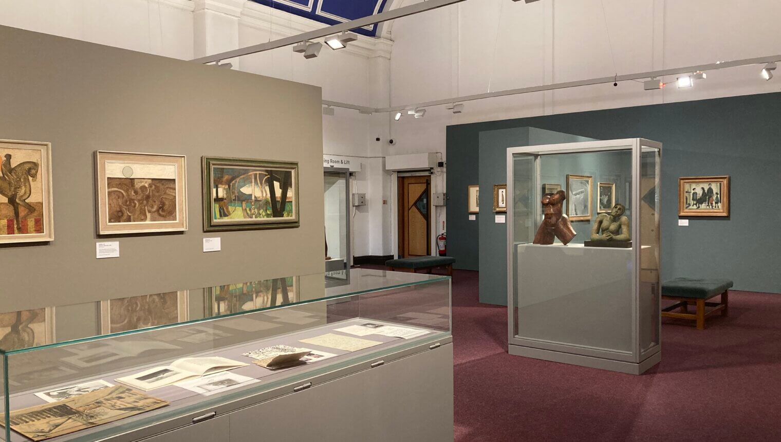 An exhibition space with a grey wall and a blue wall with paintings on them. There is also a case of paper work and a case of sculptures.