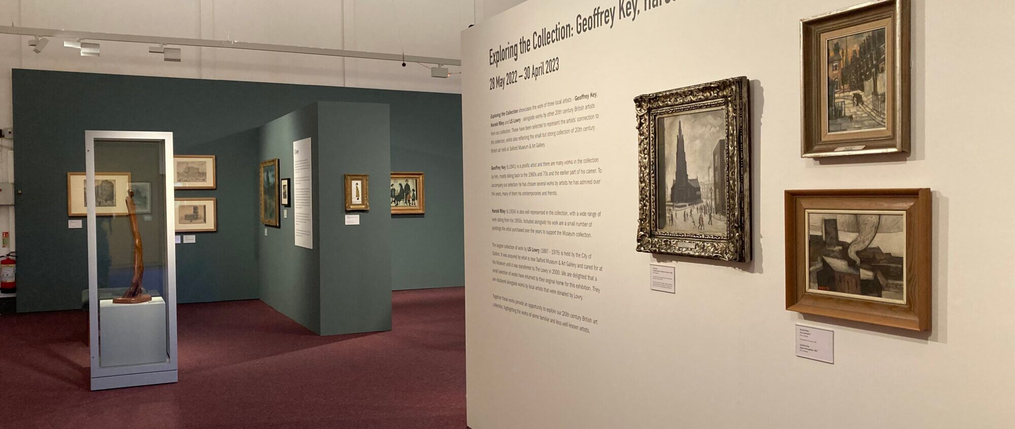 A white wall with framed paintings hanging on it and the introduction label to the exhibition. In the background are blue walls with more paintings.