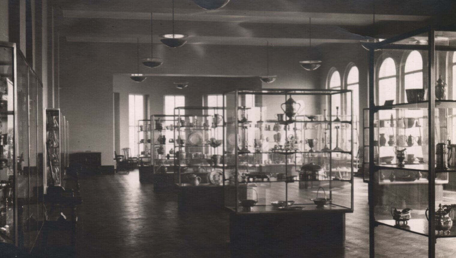 A black and white photo of a gallery with wooden floors and glass display cases full of pottery and ceramics.