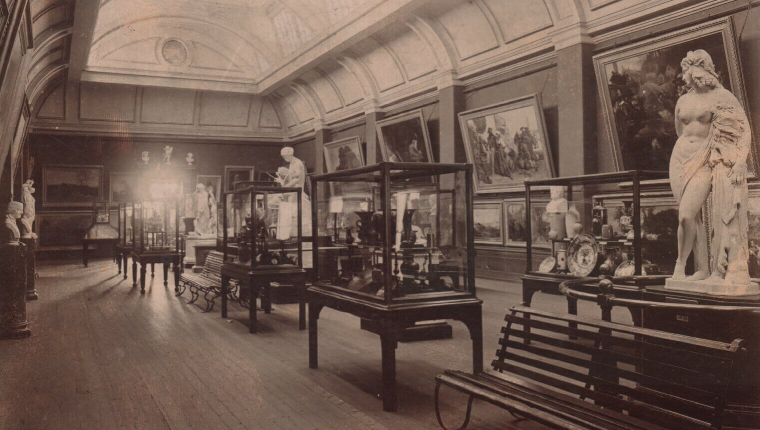 A black and white photo of a gallery with a curved ceiling. There are paintings, statues, and display cases around the room, and a wooden bench in the foreground.