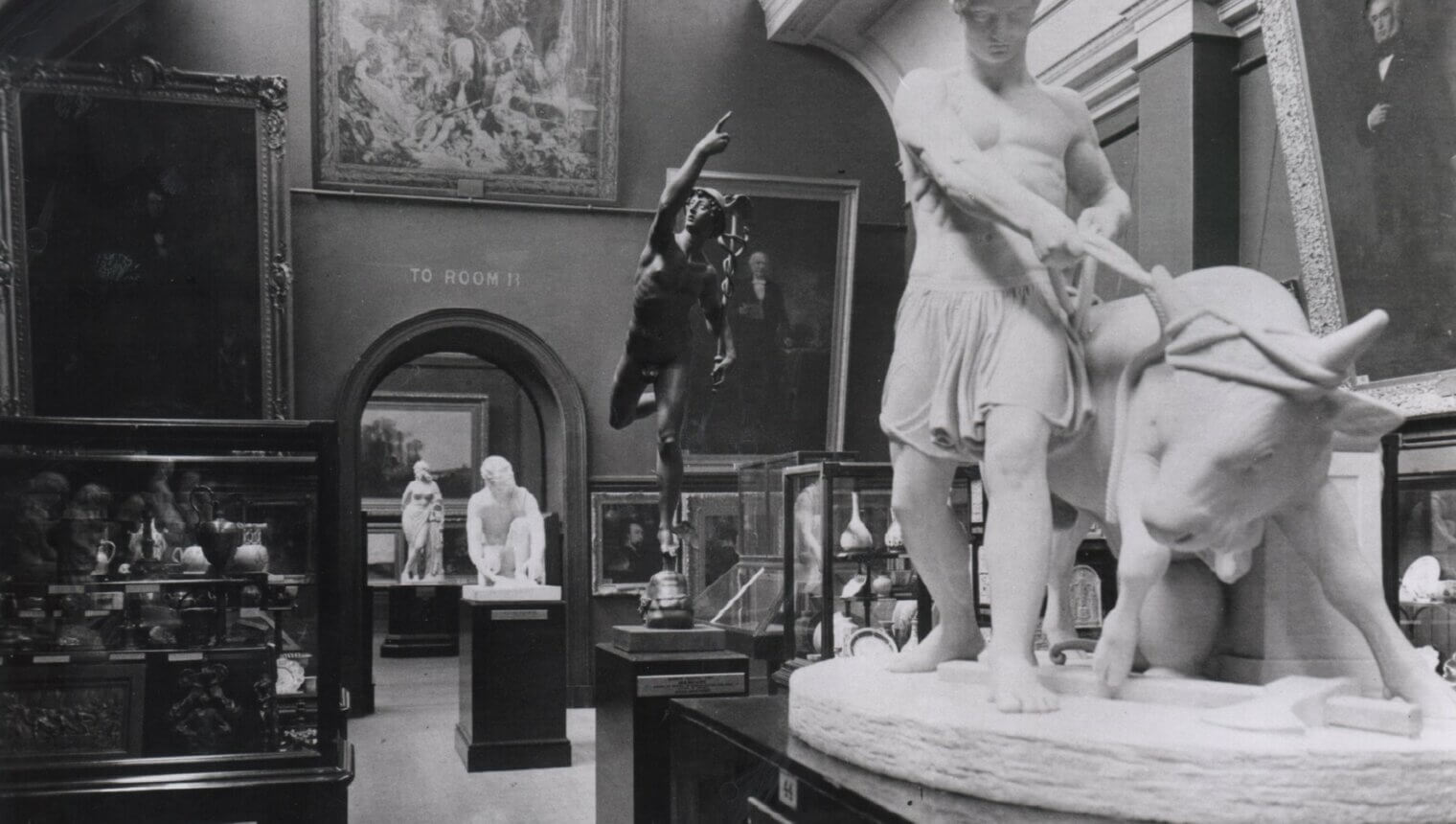 A black and white photo of a gallery. There are paintings on the wall and statues on plinths in the middle of the room. One of the statues shows a man capturing a bull.