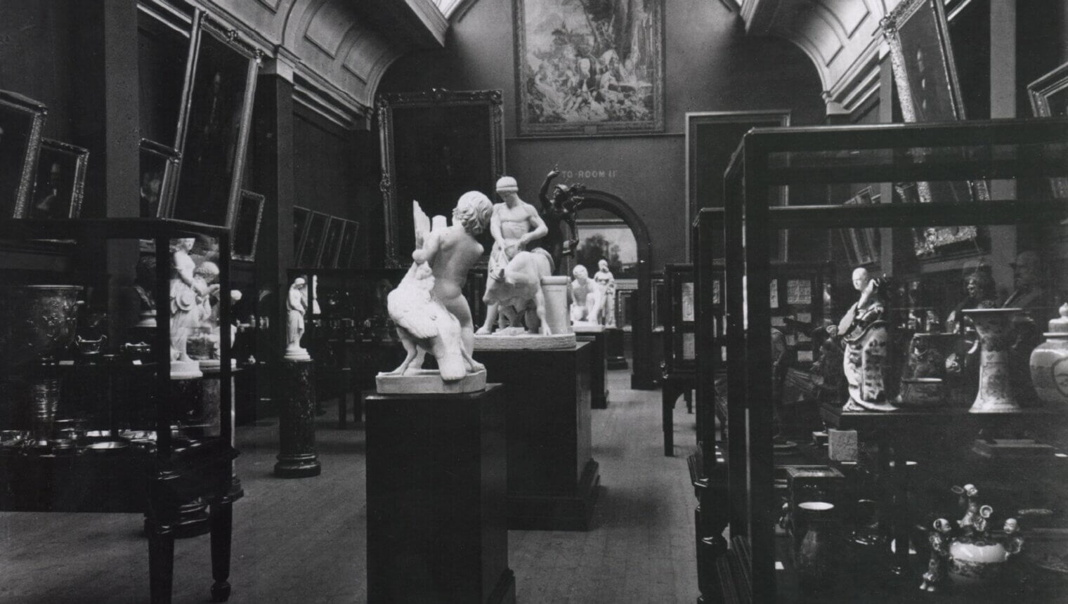 A black and white photo of a gallery space with a curved ceiling. There are marble statues on plinths in the centre of the room and display cases full of vases on either side.