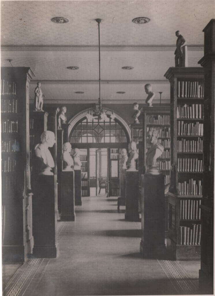 A library with aisles of books and marble busts on plinths.