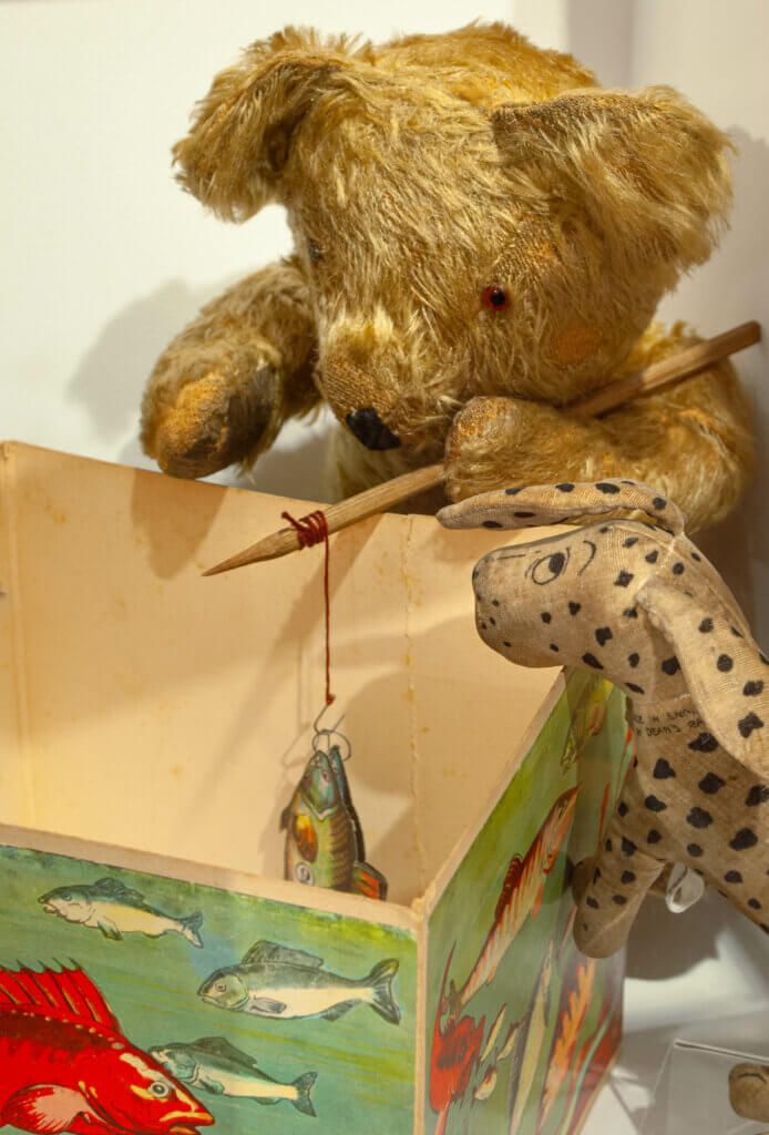 A toy dog and an old-fashioned teddy bear play a magnetic fishing game. The bear holds a fishing rod and they're both peaking into the game's box.