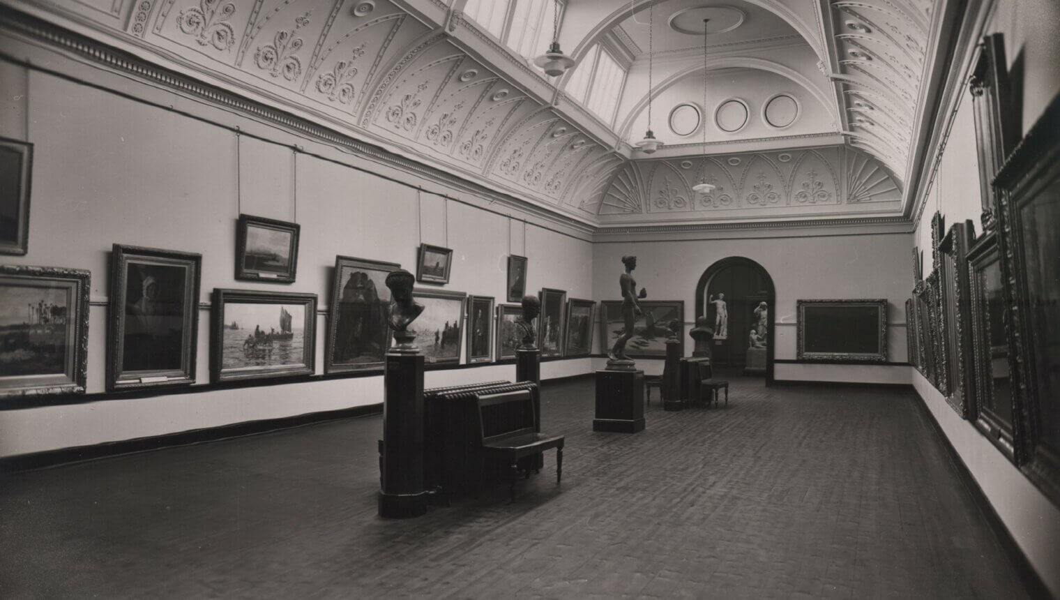 A black and white photo of a spacious gallery with a decorative, curved ceiling. There are paintings double hung on the walls, and benches in the centre of the room.