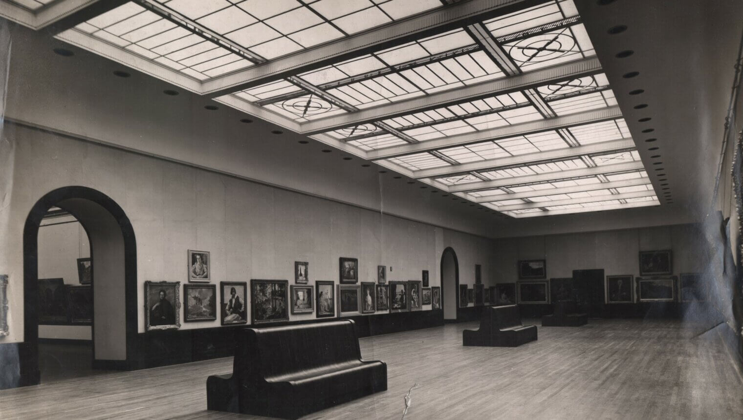 A black and white photo of a large gallery with a sky light and benches in the centre of the floor. Framed paintings hang on the walls.