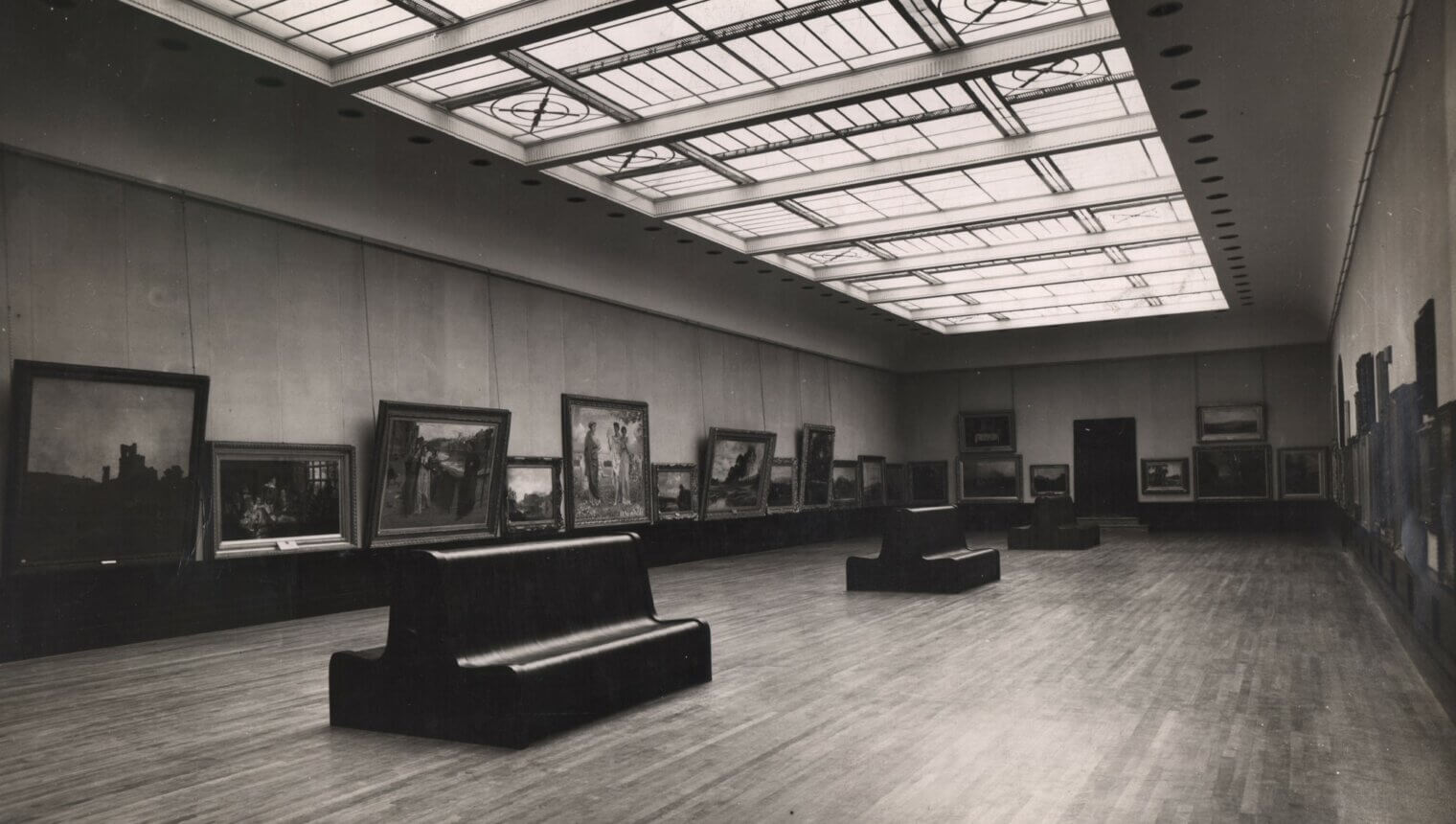 A black and white photo of a large gallery with a sky light and benches in the centre of the floor. Framed paintings hang on the walls.
