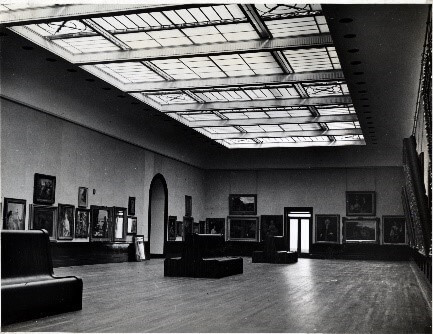 A black and white photo of a large gallery with a sky light and benches in the centre of the floor. Framed paintings hang on the walls.