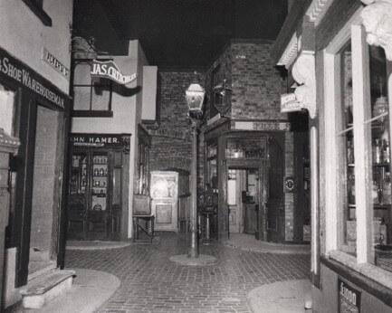 A black and white photo of a fake street with old fashioned shop fronts, cobbled floors, and a lamp post in the centre.