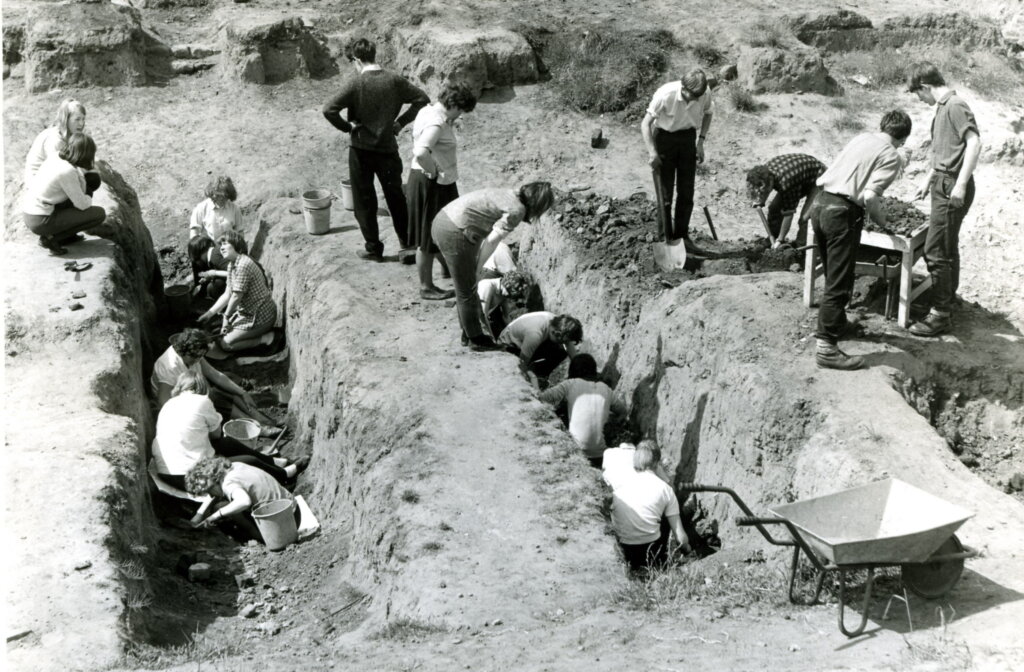 A black and white photo of an archaeological dig. People crouch in trenches in the dirt. There is a wheelbarrow in the foreground.