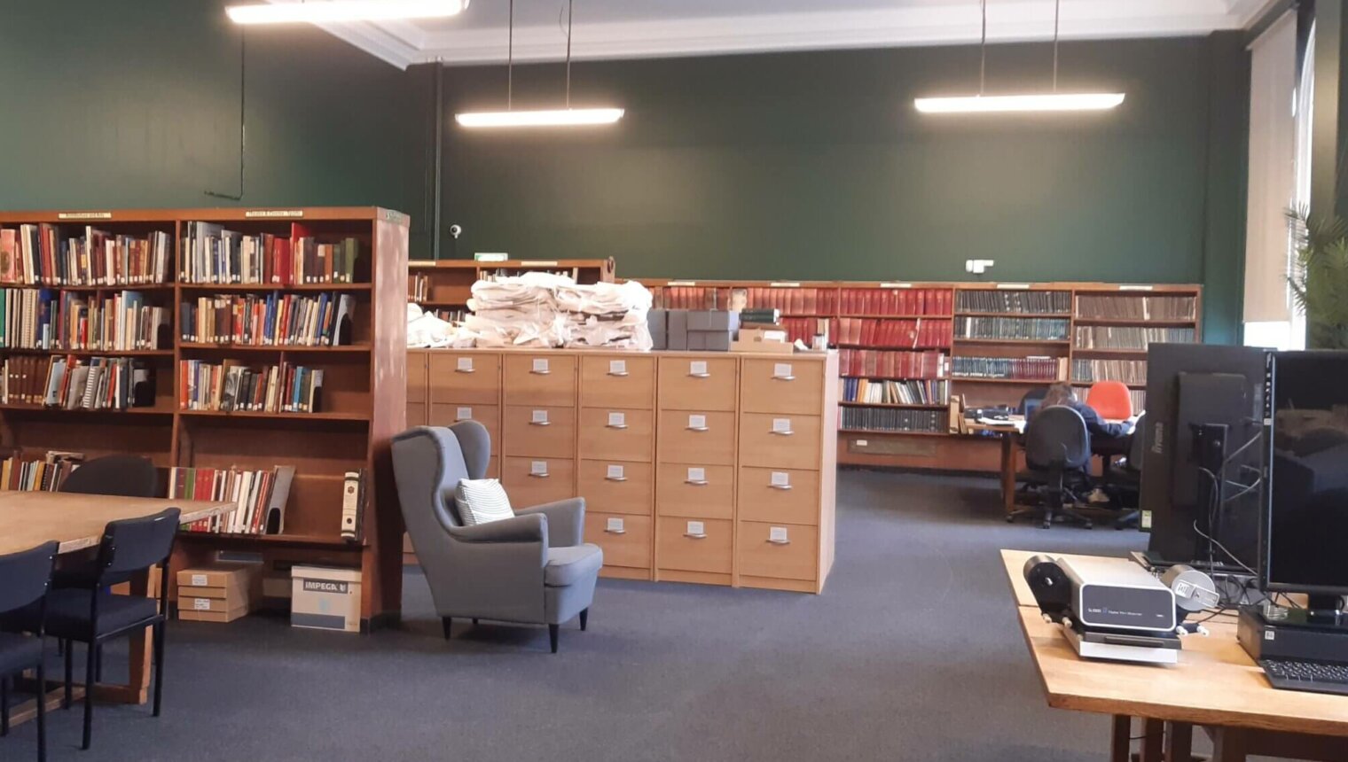 A library with green walls. There are shelves of books and computer desks. There is a blue armchair.