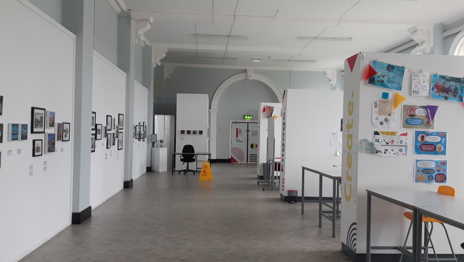 A gallery space with framed paintings hanging on the wall on the left-hand side. White studio walls and tables are on the right.