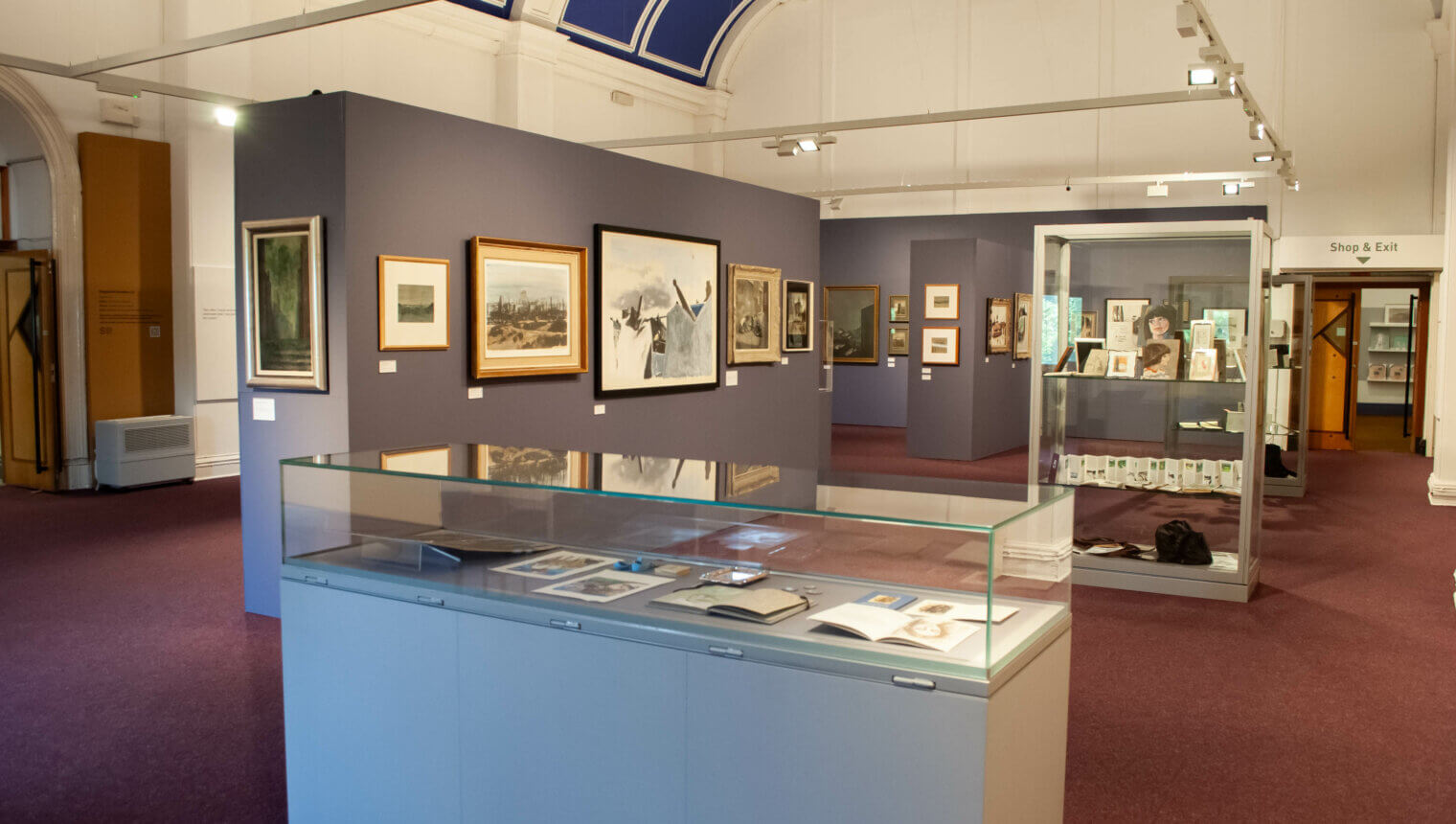 A gallery with blue walls and red carpet. There are paintings hanging on the walls and display cases holding paper.