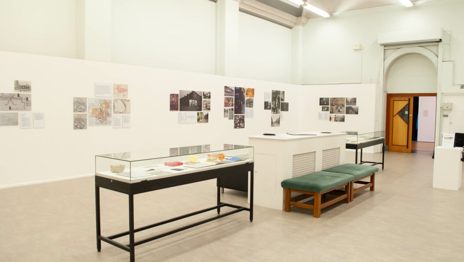 A white walled display case with images hanging on the wall. There are display cases and benches in a line in the middle of the room.