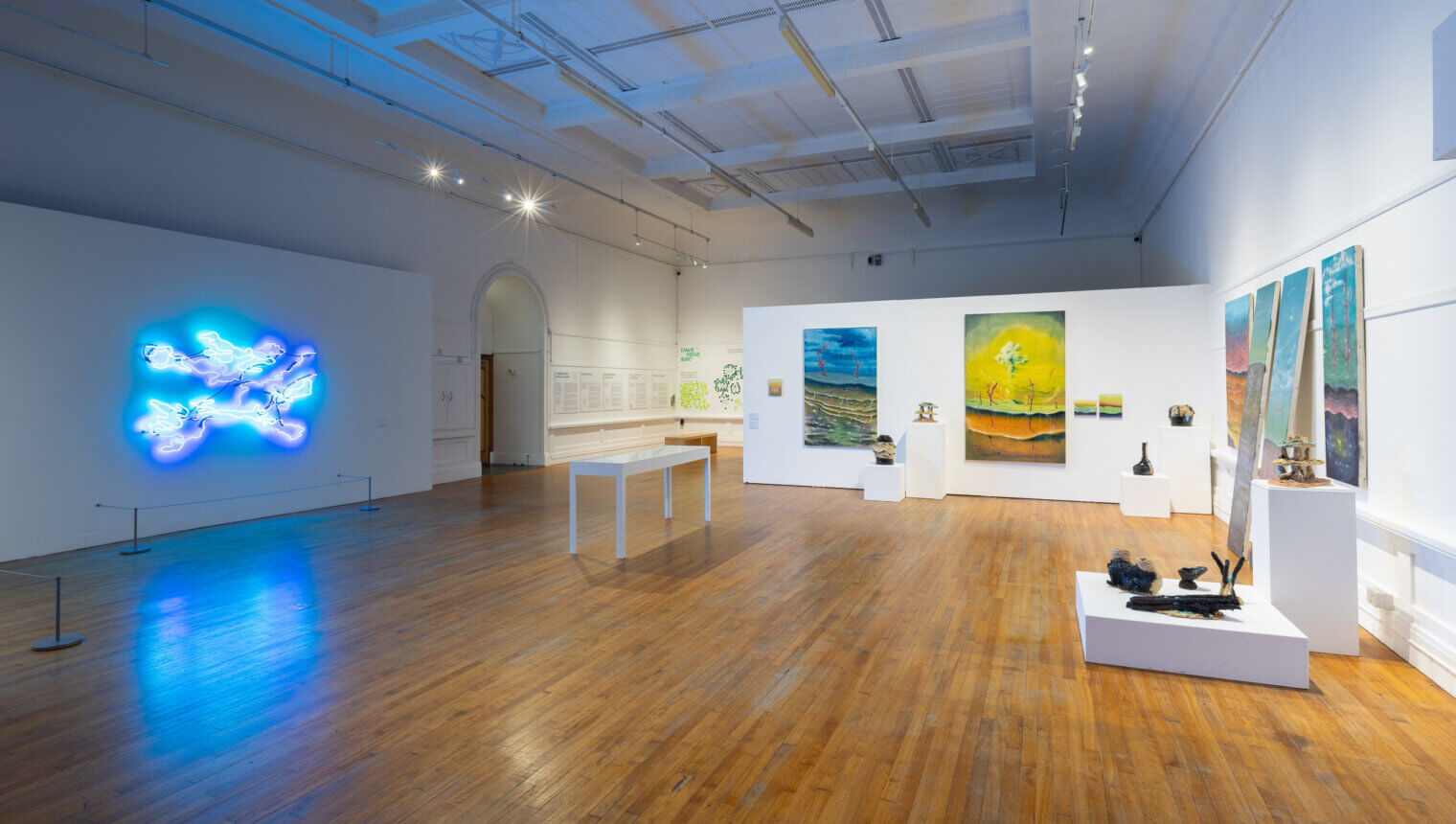 A large, white walled gallery with paintings hanging on the walls. There is an abstract artwork made from neon lights on the left-hand side.